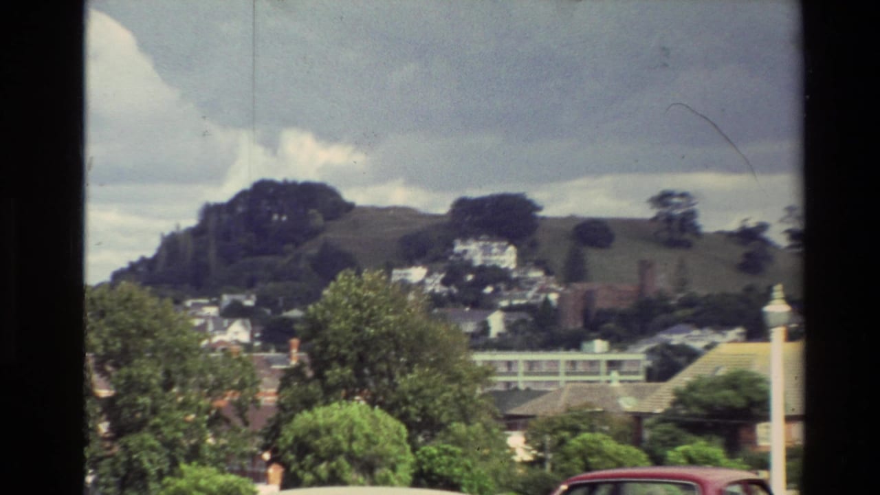 Hillside town view with trees, houses, road edge, and part of a red car.