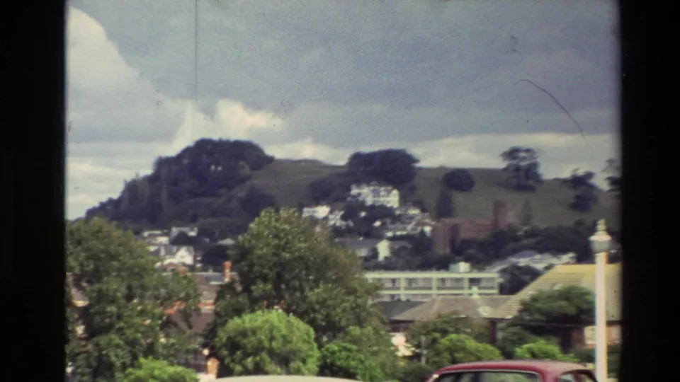 1981 New Zealand town scene with hillside homes, leafy trees, a road edge, and a red car partly visible.