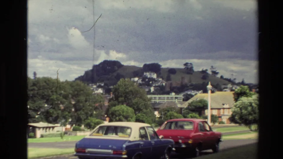 1981 New Zealand street view with a blue sedan and red car passing below hillside houses and trees.