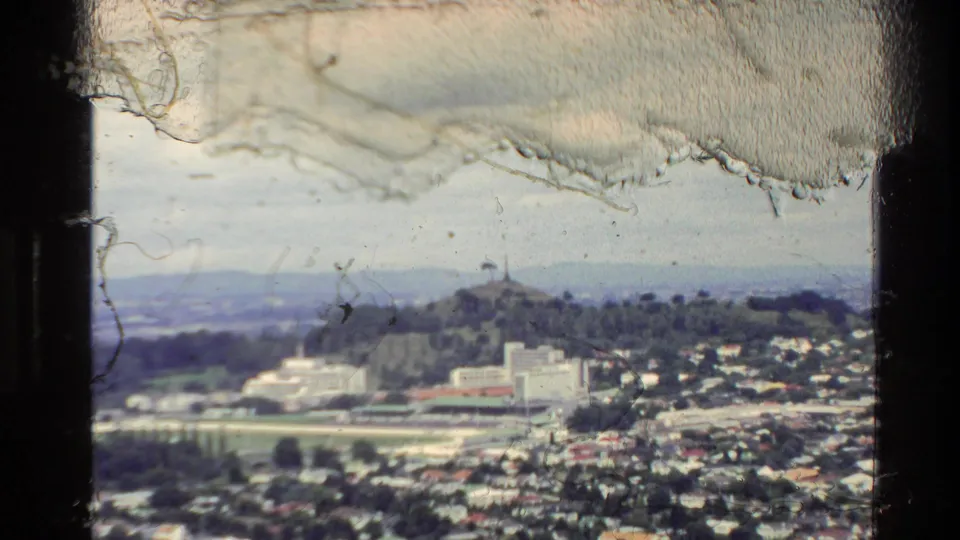 1981 New Zealand elevated city panorama with clustered houses, larger civic-looking buildings, hills, and film artifacts.
