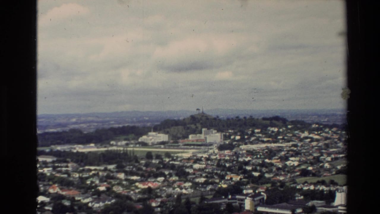 1981 New Zealand 8mm view from above, showing dense housing, a prominent hilltop structure, and distant landscape under cloud.