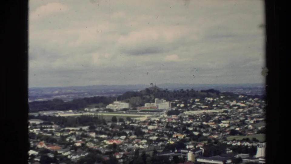 1981 New Zealand 8mm view from above, showing dense housing, a prominent hilltop structure, and distant landscape under cloud.