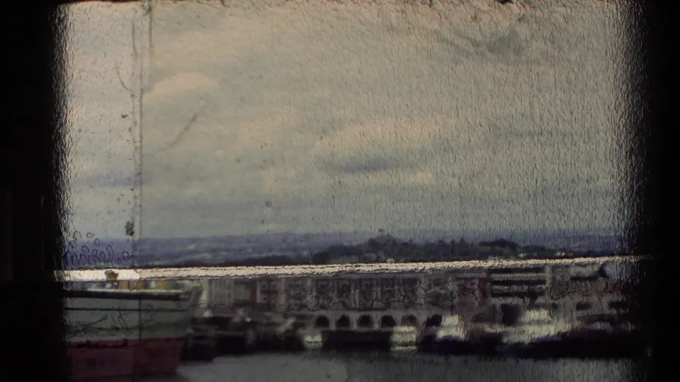 Soft 8mm frame looking across a New Zealand waterfront, with low buildings, harbor water, distant land, and heavy gate-edge shadow.