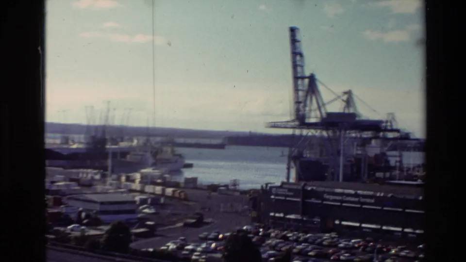 High 8mm view of a New Zealand port, with a large crane, terminal buildings, parked cars, harbor water, and visible signage.