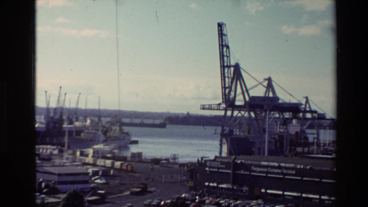 1981 New Zealand home-movie frame showing a port area with cranes, vessels, water, and industrial waterfront structures.