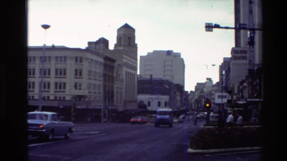 A New Zealand downtown street scene from the 1981 home movie shows traffic, older cars, multi-story buildings, and a Theatre Centre sign.