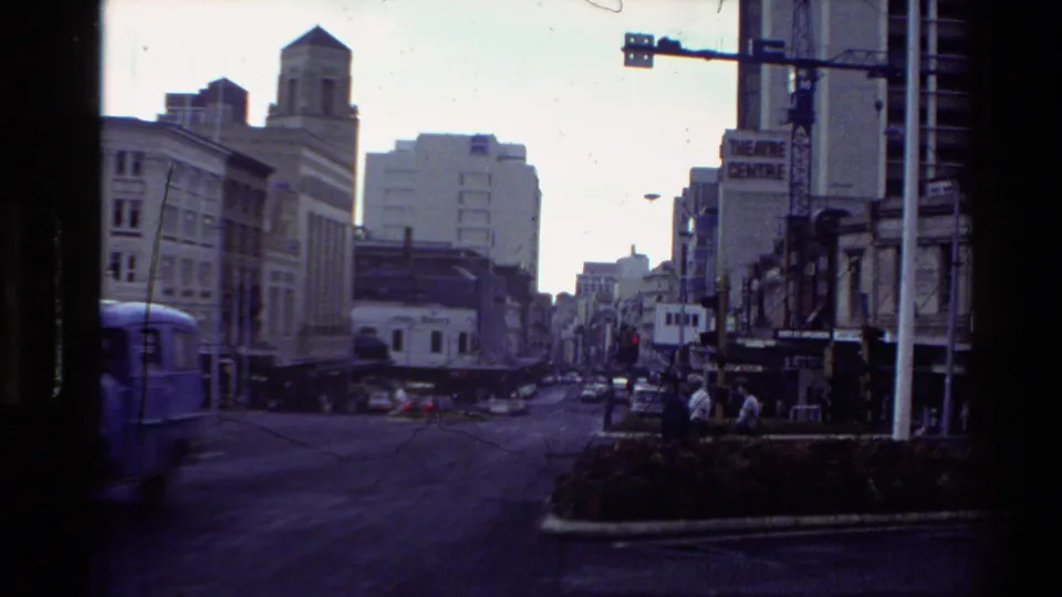 New Zealand street scene from the 1981 clip, with a blue van passing older central-city buildings and a Theatre Centre sign.