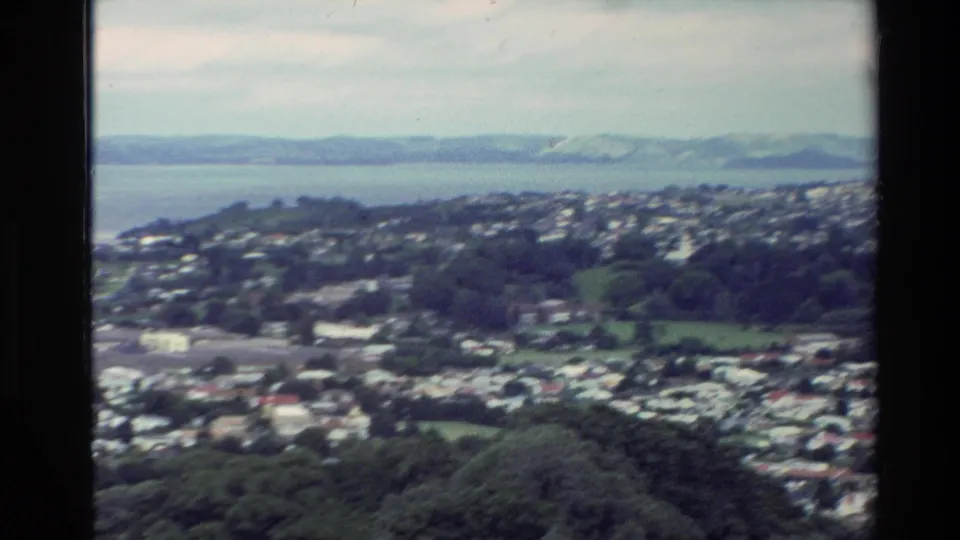 Soft 8mm frame of a New Zealand coastal city panorama, looking across hillside homes and trees toward water and far ridgelines.