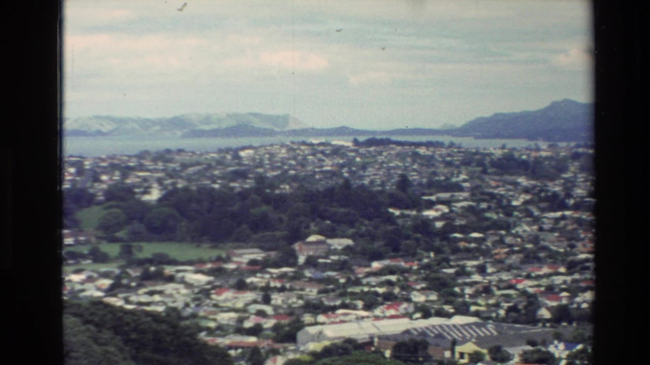 Elevated 1981 New Zealand view of a dense residential area bordered by green slopes, ocean, and distant mountain ranges.