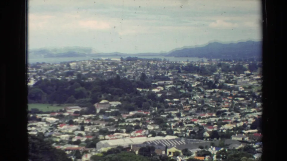 Soft 8mm frame from New Zealand in 1981 showing suburban rooftops, park-like green space, ocean, and a mountain ridge.