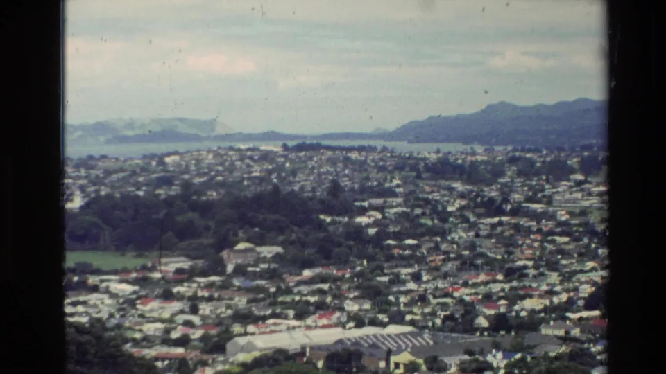 Final 1981 New Zealand panorama frame with a bird in the sky above town rooftops, ocean water, and distant mountains.