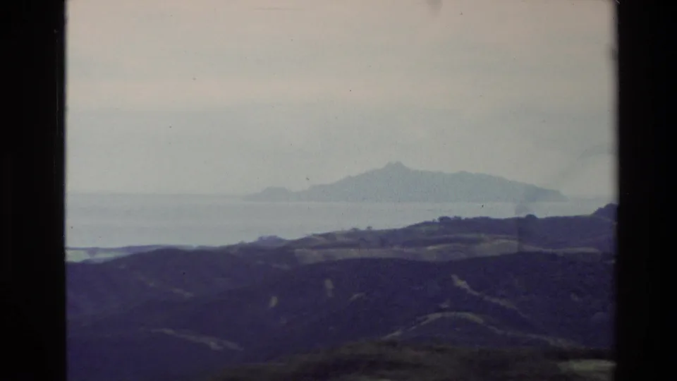1981 New Zealand home-movie frame showing rolling coastal hills, open water, and a distant mountain-like island.