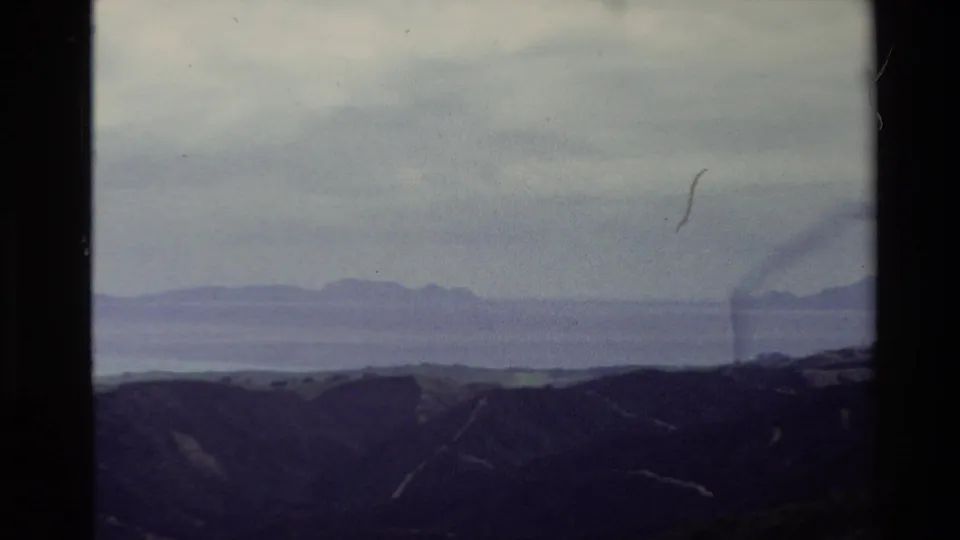 1981 New Zealand 8mm landscape frame with low foreground hills, pale sea, and distant landforms on the horizon.