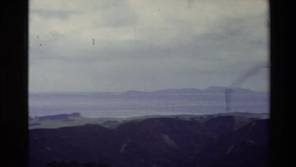 1981 New Zealand home-movie still showing hill ridges, a broad waterway, and distant low islands in hazy light.