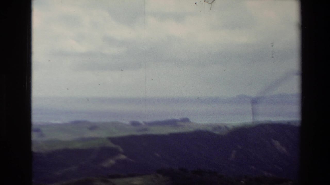 1981 New Zealand 8mm frame showing a hazy coastal lookout view with rolling hills, water, distant landforms, and cloudy sky.