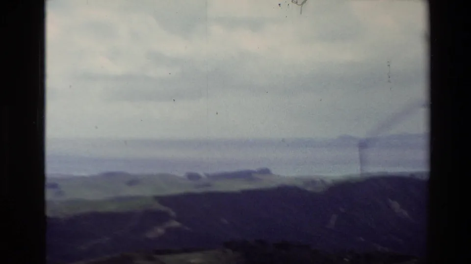 1981 New Zealand 8mm frame showing a hazy coastal lookout view with rolling hills, water, distant landforms, and cloudy sky.