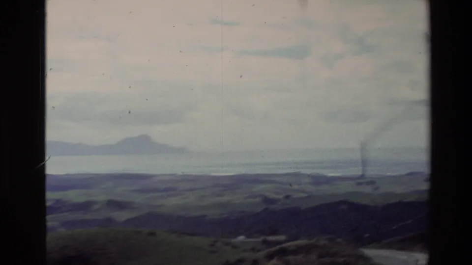 1981 New Zealand home-movie frame with green hills in the foreground, water beyond, distant island shapes, and visible 8mm frame marks.