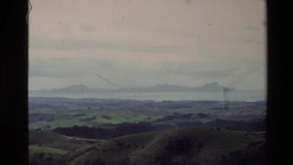 1981 New Zealand 8mm view across rounded green hills toward a broad body of water and hazy distant land under cloud.
