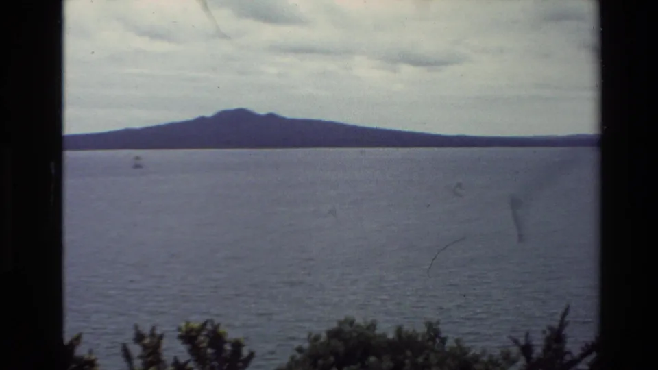 1981 New Zealand 8mm frame showing a dark island or mountain profile across water, with low clouds and foreground shrubs.