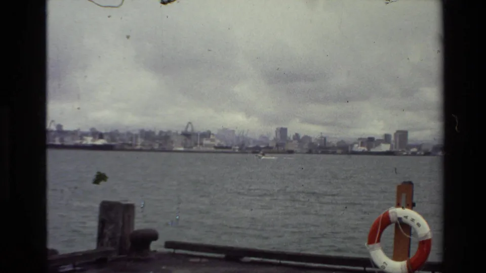 1981 New Zealand dockside view across a wide harbor or river toward a distant city skyline, with a life ring in the foreground.