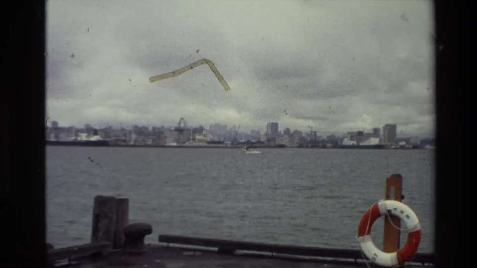 A 1981 New Zealand waterfront view from a dock, looking across open water to high-rises and cranes beneath an overcast sky.