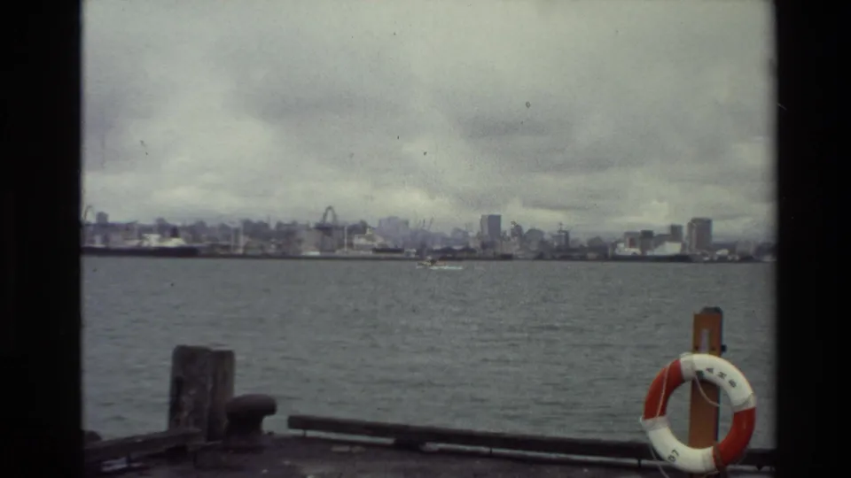 Soft 8mm frame of a New Zealand harbor or river view in 1981, with a skyline, cranes, and dockside safety ring visible.