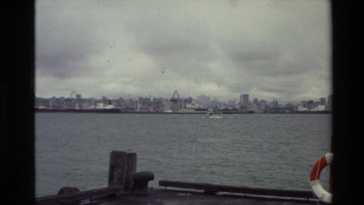 1981 New Zealand 8mm boat-view frame looking across water toward a distant working waterfront under cloudy sky.