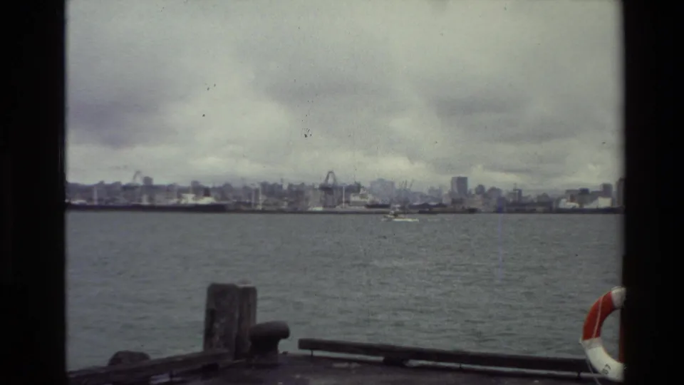1981 New Zealand 8mm boat-view frame looking across water toward a distant working waterfront under cloudy sky.