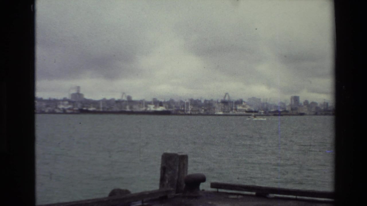Distant harbor skyline across water with a wooden post and pier edge in foreground.