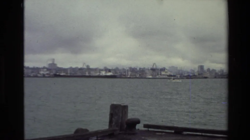 1981 New Zealand harbor view from a boat, showing open water, a low skyline, cranes, and foreground dock hardware.