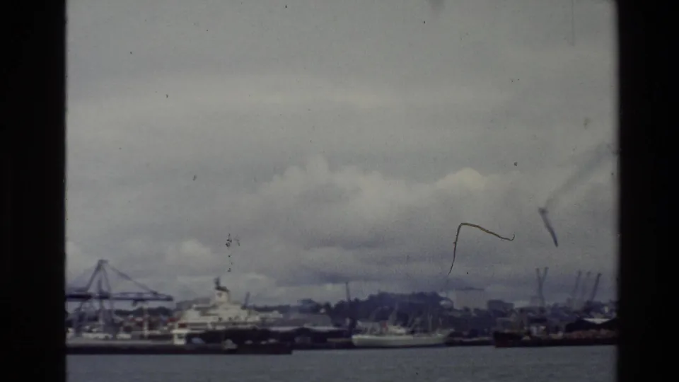 1981 New Zealand waterfront frame with moored boats and cranes seen across the water through scratched 8mm film.