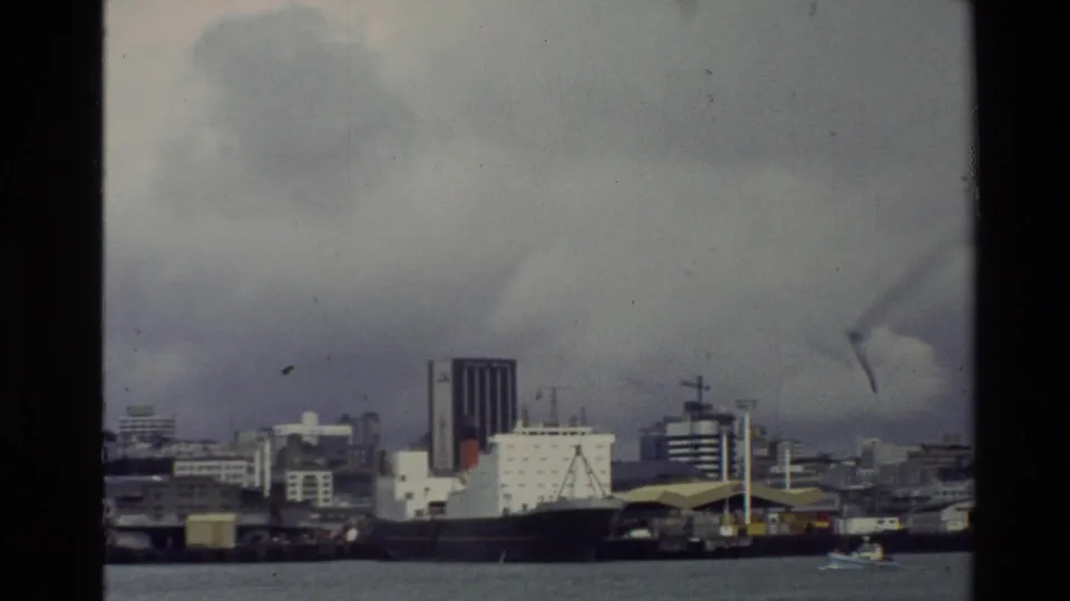 1981 New Zealand 8mm view of a docked cargo ship, waterfront buildings, gray water, and overcast harbor sky.