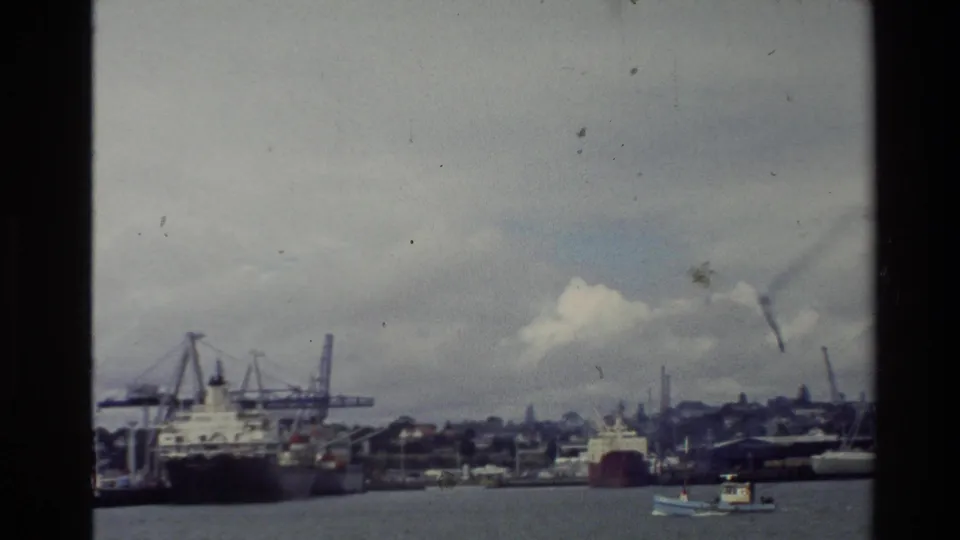 1981 New Zealand port view with cranes, docked vessels, low shoreline structures, gray harbor water, and a small boat in the foreground.