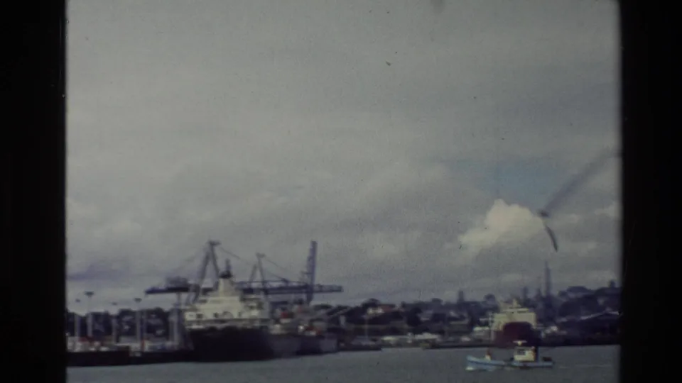 Vintage 8mm harbor frame from New Zealand showing cranes, moored ships, a small boat, distant shoreline, and a cloudy sky.