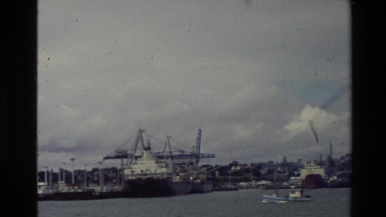 1981 New Zealand 8mm view of a working harbor with cargo vessels, cranes, water, and cloudy hills in the distance.