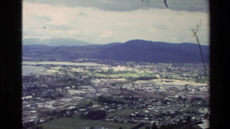 Elevated 8mm view in New Zealand, 1981, with a town, shoreline, open fields, bay water, and distant hills.