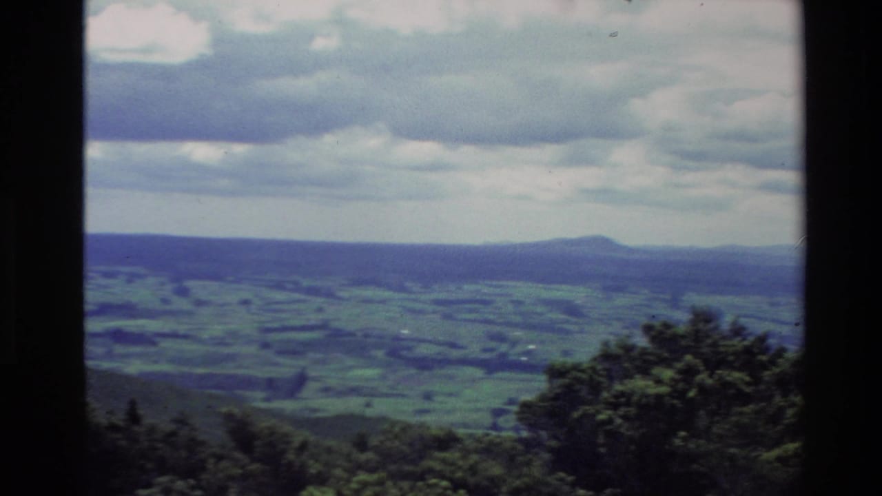 Wide countryside view with green fields, low hills, trees, and a cloudy horizon.