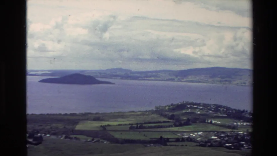 1981 New Zealand 8mm frame showing a coastal vista with an offshore island, broad water, patchwork farmland, and distant hills.