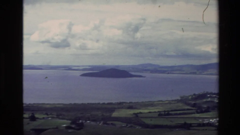1981 New Zealand home-movie frame of an offshore island and wide water view, with rural fields below and slight handheld blur.