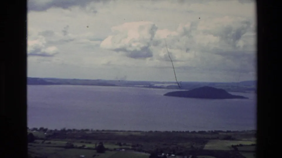 1981 New Zealand home-movie view across open coastal water toward a small island and distant low hills under broken cloud.