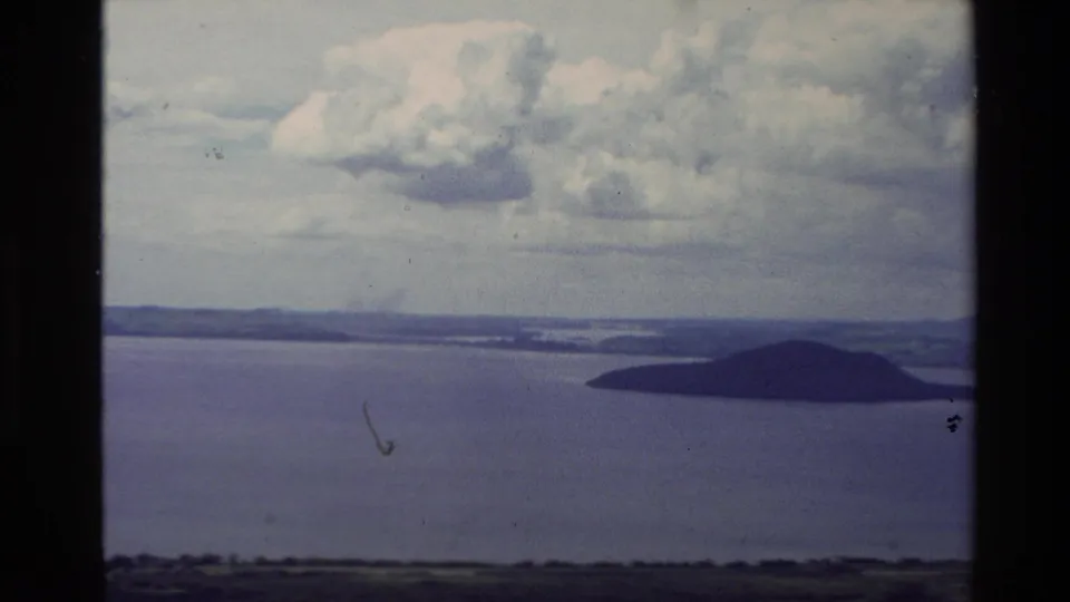 Elevated 1981 New Zealand coastal frame showing a small offshore island beyond rural fields and still bay water.