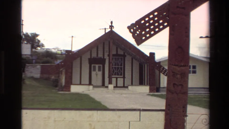 1981 New Zealand 8mm frame showing a gabled timber building with carved details, a path, grass, and a carved post in the foreground.