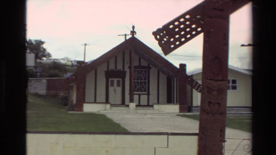 Home-movie frame from New Zealand showing a carved foreground post and a gabled building with decorative timberwork across a yard.