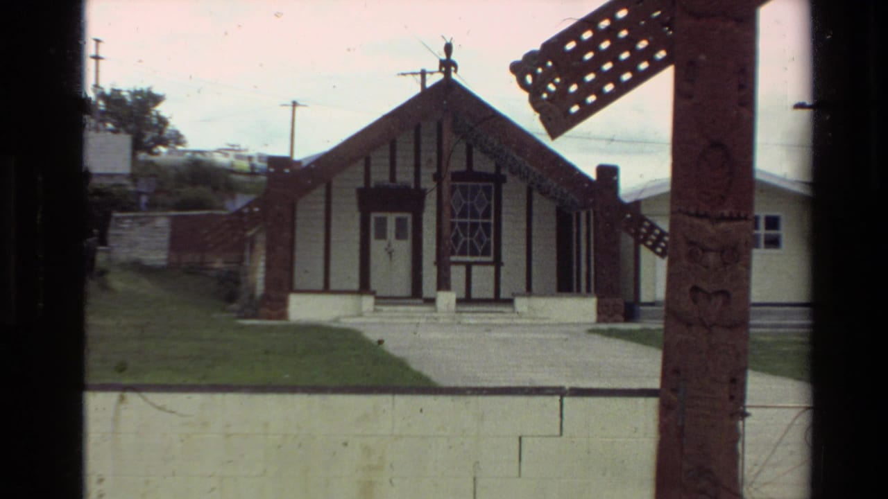 1981 New Zealand home-movie frame showing a wharenui-style building with white walls, dark timber trim, lawn, and carved foreground post.