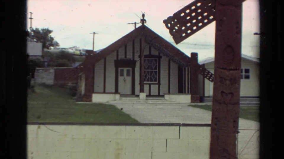 1981 New Zealand home-movie frame showing a wharenui-style building with white walls, dark timber trim, lawn, and carved foreground post.