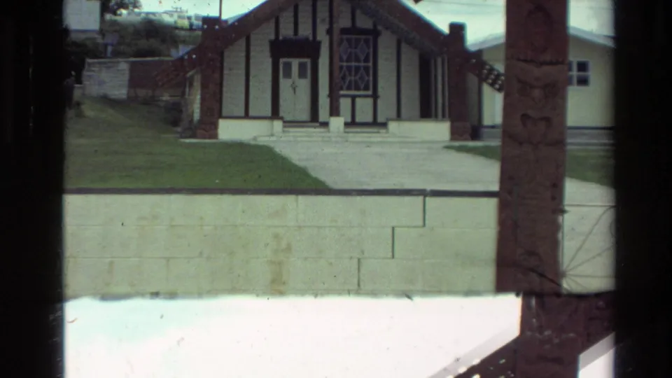 1981 New Zealand 8mm frame of a wharenui-style exterior, with dark trim, pale retaining wall, lawn, and carved post near the right edge.