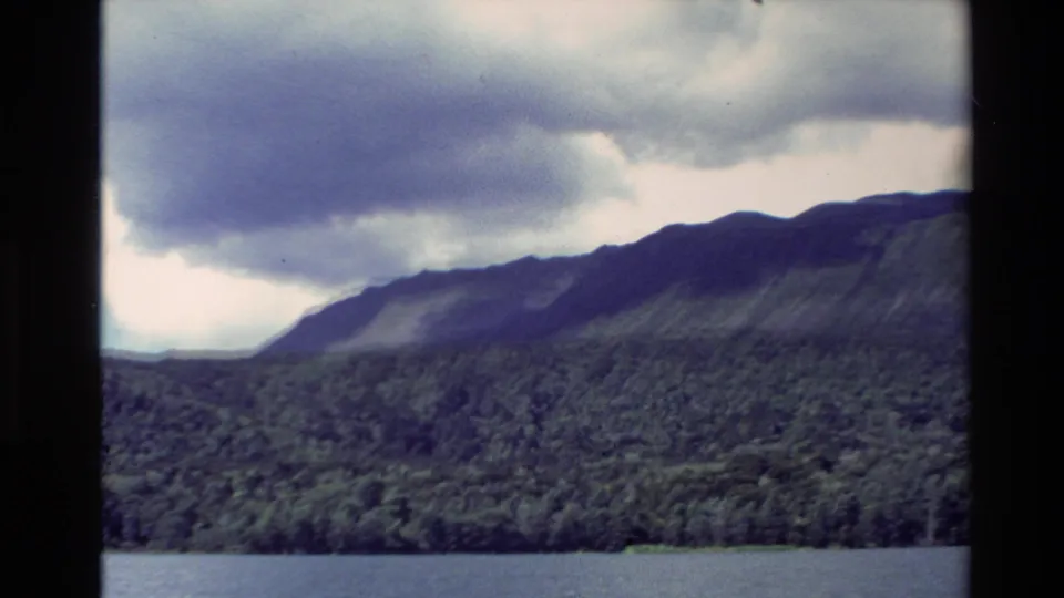 1981 New Zealand 8mm frame of a forested shoreline and mountain slopes across calm water under a heavy cloud bank.