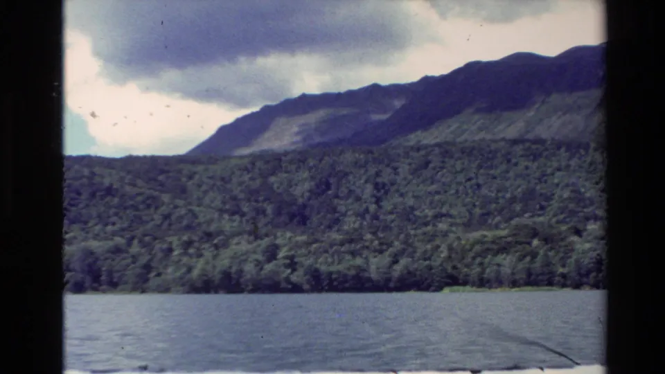 1981 New Zealand 8mm frame showing a lake, dense green shoreline, and dark mountain ridge beneath heavy clouds.
