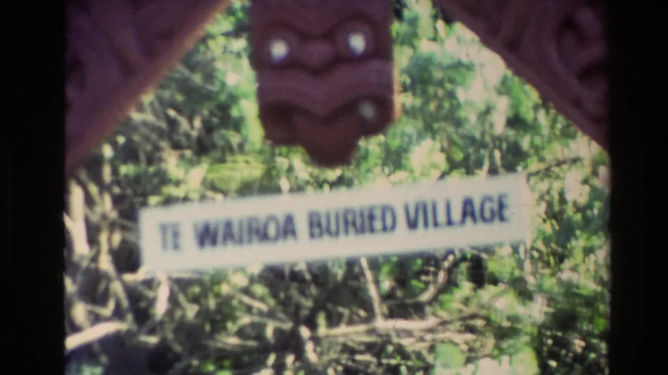 1981 New Zealand 8mm frame with a sign reading Te Wairoa Buried Village, seen through dark foreground shapes and surrounding greenery.