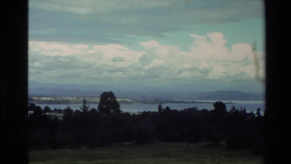 1981 New Zealand travel frame showing a wide lake view with dark foreground trees, distant hills and layered cloud.
